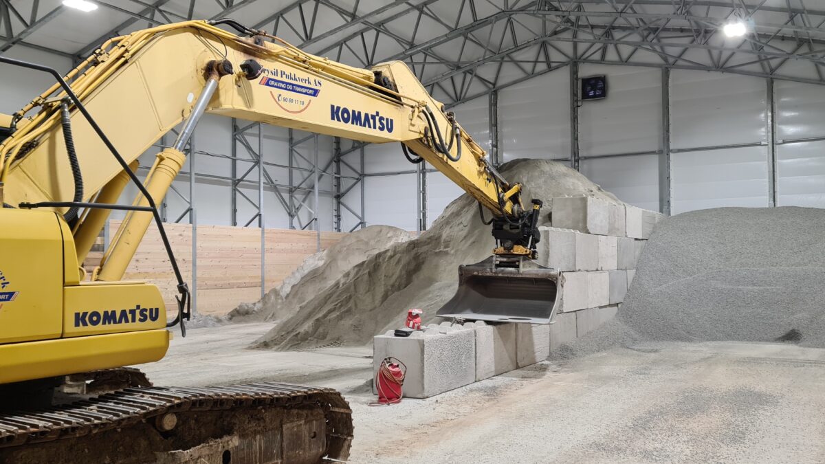 En gul grävmaskin inuti en bulkhall med högt tak, hanterar stora betongblock och sand. Hallgruppen har byggt denna robusta struktur.