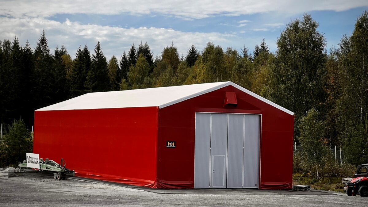 En röd bulkhall med en vit tak, omgiven av skog. Hallgruppen tillhandahåller denna flexibla och hållbara lösning för olika förvaringsbehov.