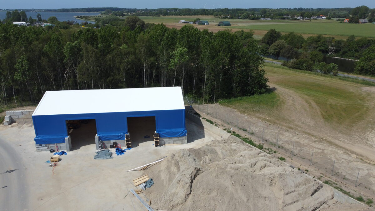 Hallgruppen har byggt en blå bulkhall vid vattnet, omgiven av grönska. Perfekt för förvaring och logistik nära hamnområden.