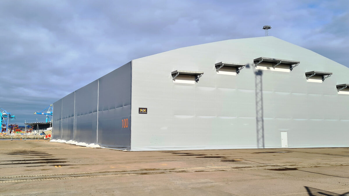En grå bulkhall vid en hamn med kranar i bakgrunden, byggd av Hallgruppen. Robust konstruktion för maritima miljöer.