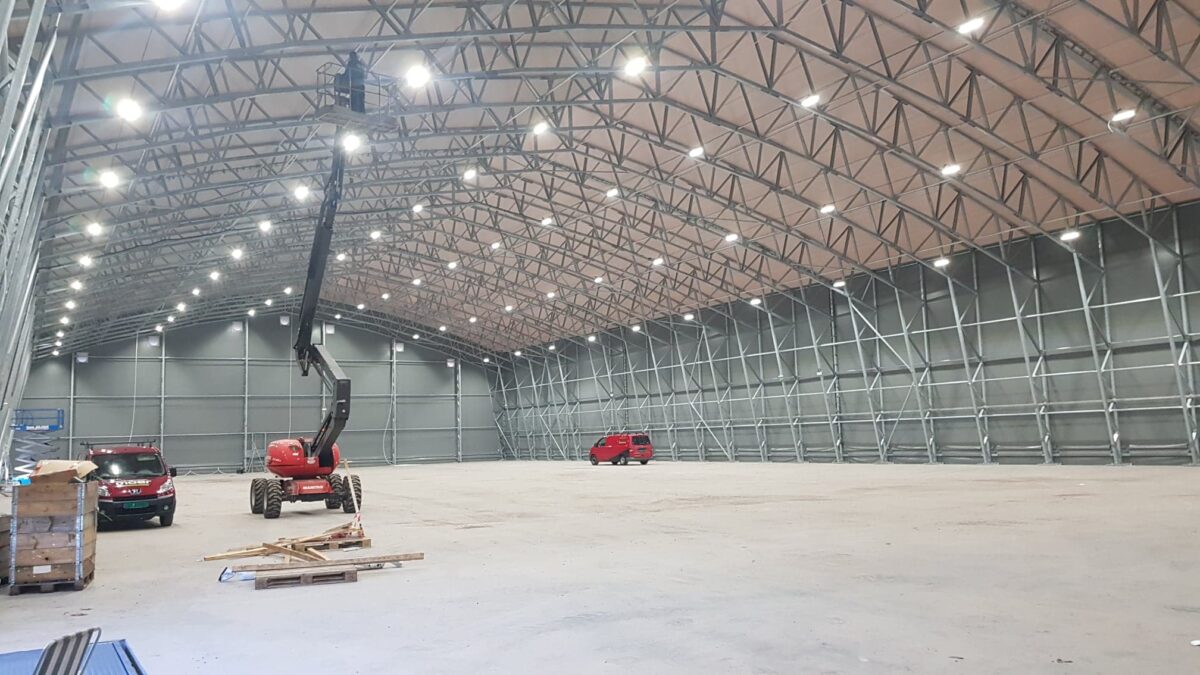 Interiören av en rymlig lagerhall med stålkonstruktion, en lyftkran och två röda fordon. Byggnaden är uppförd av Hallgruppen.