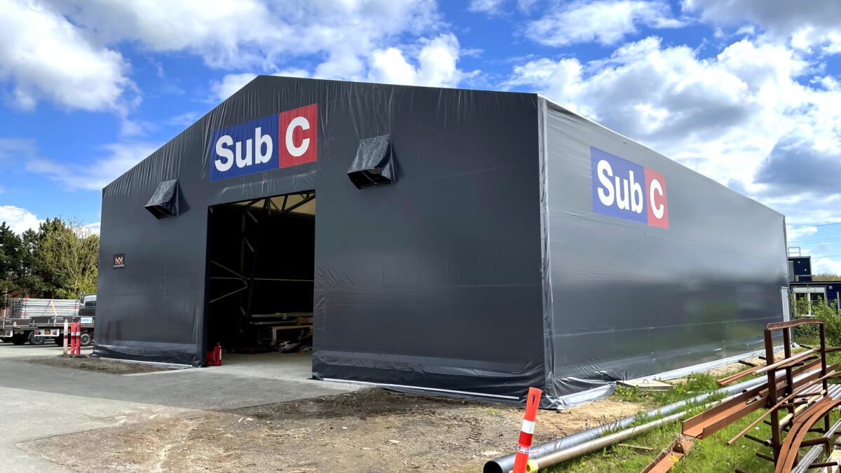 En grå lagerhall från Hallgruppen med logotypen 'Sub C', omgiven av industriell utrustning.