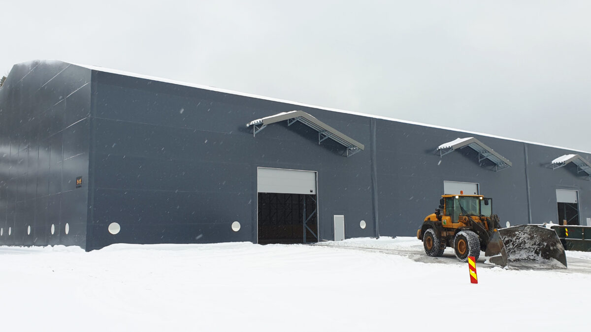 En stor grå lagerhall från Hallgruppen under snöfall, med arbetsfordon framför.