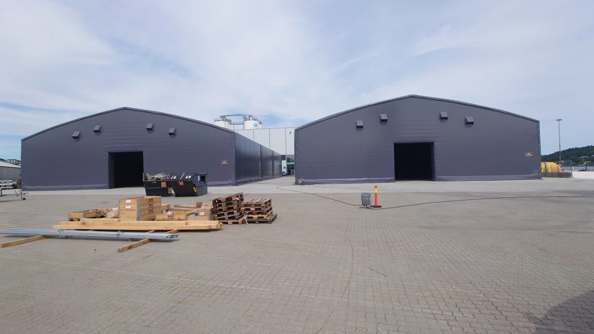 Två stora svarta lagerhallar på en industriell arbetsplats, byggda av Hallgruppen, med pallar och lastmaterial framför.