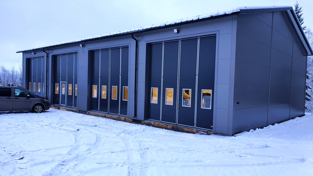 En grå maskinhall med stora glasfönster, belägen i ett snötäckt område, en bil parkerad bredvid.