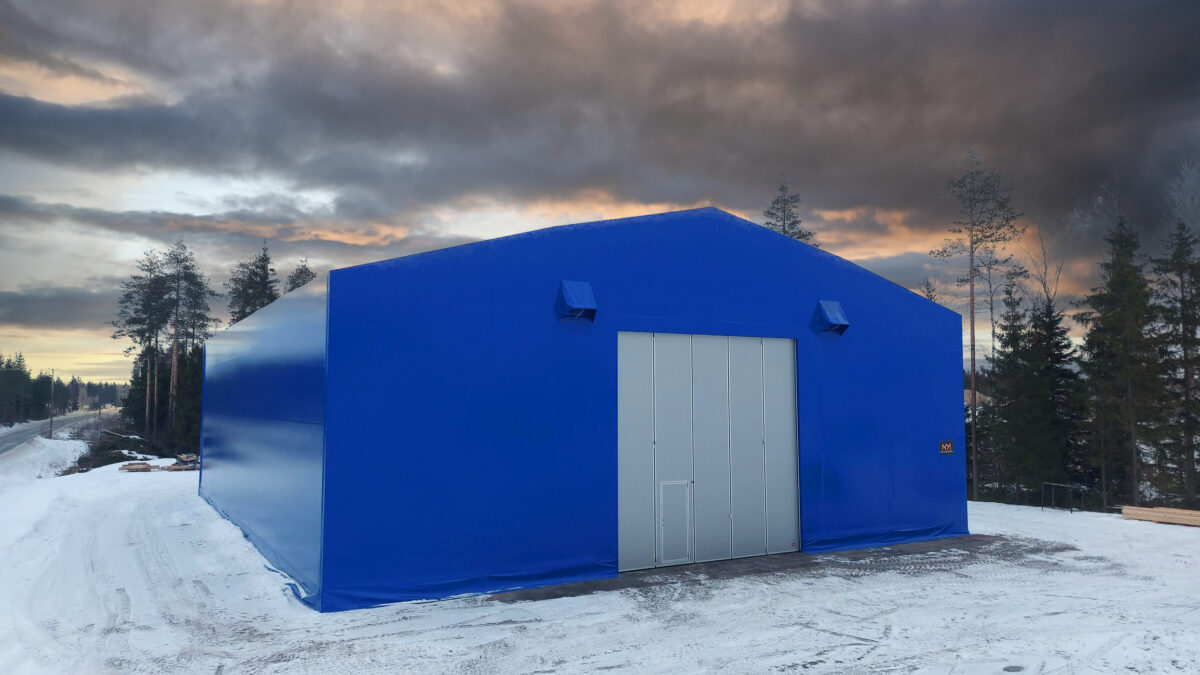 En blå maskinhall i ett snötäckt skogslandskap, under en dramatisk himmel med solnedgång.