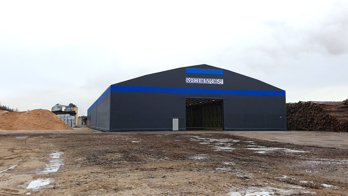 Ett grått lager med Moelven-logotypen, uppfört av Hallgruppen, står på ett industriområde. Perfekt för träförvaring med stora staplar av sågspån och timmer vid sidan.