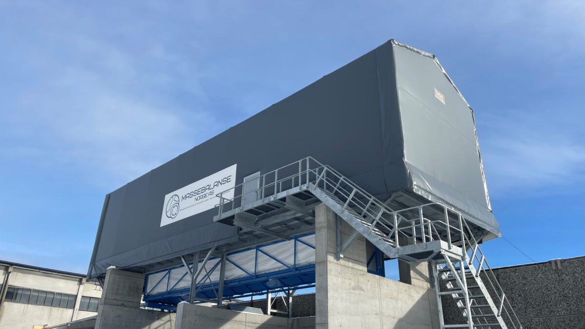 En hög, grå täIthall från Hallgruppen står på en upphöjd plattform med en klarblå himmel i bakgrunden.