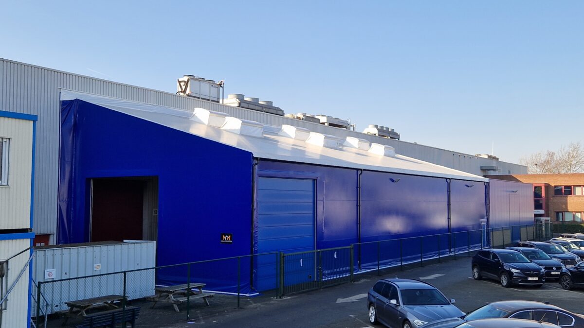 En blå täIthall byggd av Hallgruppen står vid en industriell byggnad, med parkerade bilar framför.