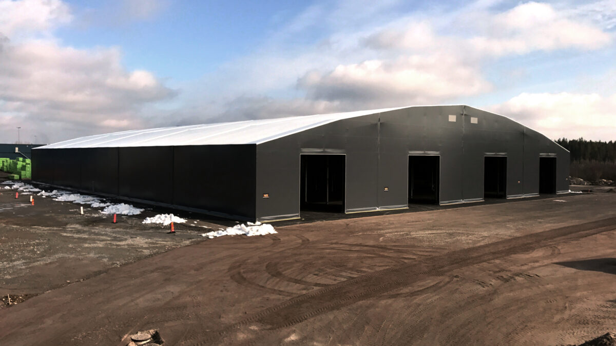 En stor svart täIthall uppförd av Hallgruppen står på en byggarbetsplats under en klar himmel.
