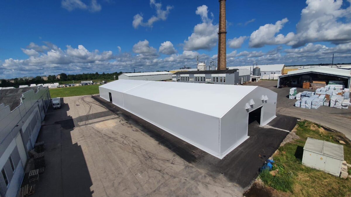 En stor vit täIthall byggd av Hallgruppen står på en industriell plats med gröna fält i bakgrunden.