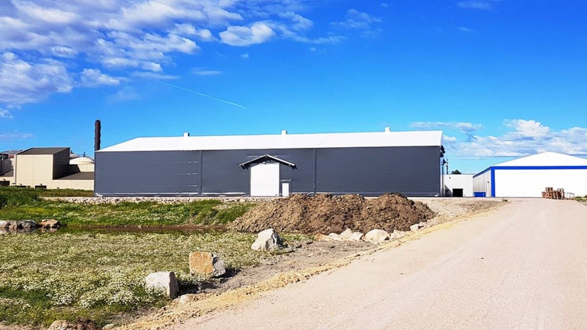 En grå täIthall byggd av Hallgruppen står på en öppen yta, omgiven av gröna fält och andra byggnader.