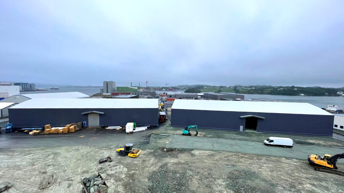 En stor grå täIthall från Hallgruppen står på en byggarbetsplats nära en hamn, omgiven av byggfordon.