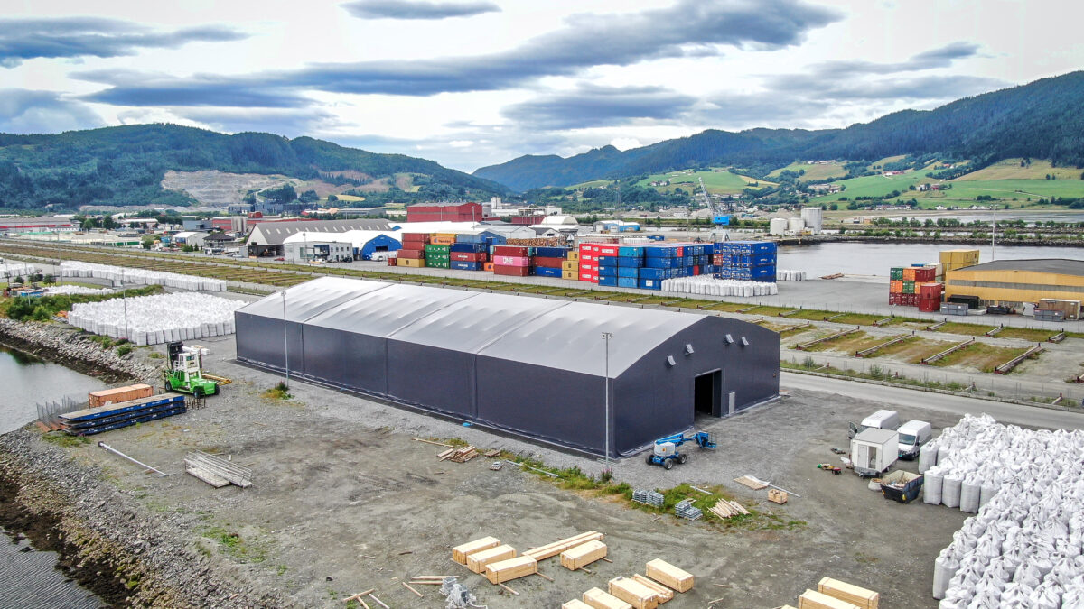 Hallgruppen har byggt en grå täIthall nära en hamn, med färgglada container i bakgrunden.