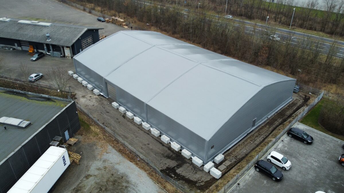 En grå täIthall uppförd av Hallgruppen står på en industriell plats med parkerade bilar och byggnader i närheten.